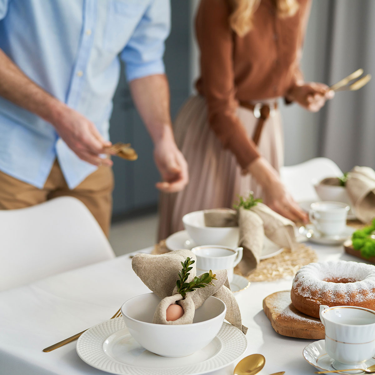 Due persone stanno preparando un tavolo imbandito per un pranzo elegante, con stoviglie e decorazioni naturali. Una tavola adornata con tovaglioli piegati e un dolce al centro.