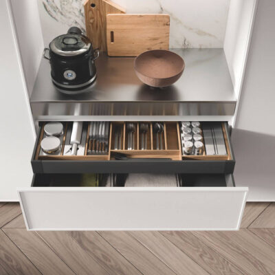 Immagine di un cassetto della cucina aperto, organizzato con posate in legno e contenitori bianchi, un recipiente in ceramica e un riscaldatore elettrico sullo sfondo.