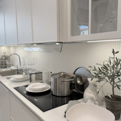 Interno di una cucina moderna con ripiani bianchi, stoviglie eleganti, pentole in acciaio inox e piante decorative sul tavolo.