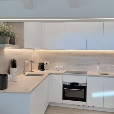 Cucina moderna con mobili bianchi, piano in legno chiaro e illuminazione a LED, dotata di lavello, forno e accessori.