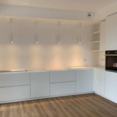 Cucina moderna con mobili bianchi e lineari, illuminazione a sospensione, pavimento in legno e nuovi elettrodomestici.