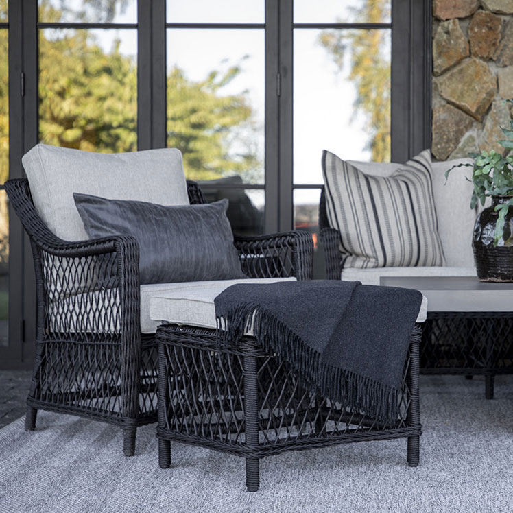 Poltroncina in rattan nero con cuscini chiari e plaid scuro, posizionata in un ambiente luminoso e accogliente.