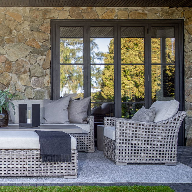 Un'area esterna elegante con divani in rattan e cuscini morbidi, circondata da pareti in pietra e finestre che riflettono la natura.