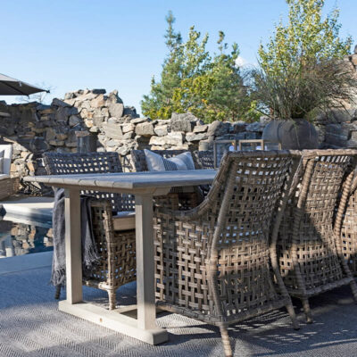 Un elegante tavolo in legno circondato da sedie in vimini, situato vicino a una piscina, con un ombrellone e una vista su rocce naturali e alberi.