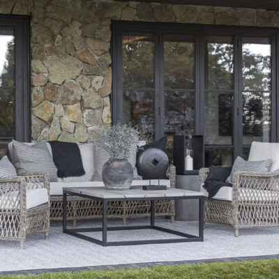 Un accogliente spazio esterno arredato con un divano in rattan, cuscini decorativi e un tavolino moderno, circondato da pareti in pietra e finestre larghe.