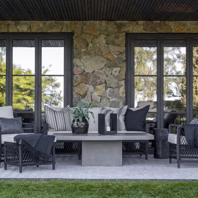 Spazio esterno elegante con divani e poltrone in rattan nero, tavolino moderno e decorazioni minimaliste, circondato da finestre in legno.