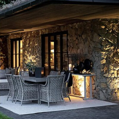 Un accogliente patio esterno con arredamento in rattan, illuminato da luci soffuse, circondato da una parete in pietra e piante decorativi.