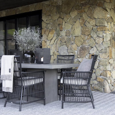 Una elegante spazio esterno con un tavolo grigio e sedie in rattan, circondata da una parete di pietra e decorazioni verdi.