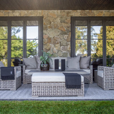 Un accogliente salotto esterno con divano e poltrone in rattan intrecciato, cuscini chiari, tavolino centrale e un tappeto sul prato verde.