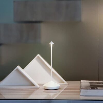 Lampada moderna con base geometrica, simile a una piramide, posizionata su un tavolo accanto a oggetti decorativi. Sfondo minimalista.