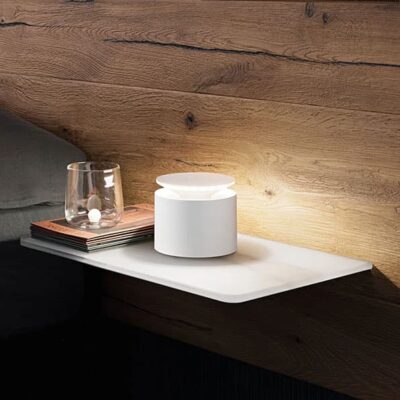 Una lampada moderna bianca su un comodino in legno, accanto a un bicchiere e una pila di riviste. Un'atmosfera calda e accogliente.