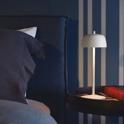 Una lampada elegante su un tavolino accanto a un letto, con lenzuola scure e un libro rosso sul tavolino. L'atmosfera è rilassante e moderna.