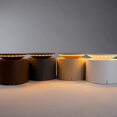 Quattro lampade moderne con base cilindrica in diverse tonalità, illuminate con luce soffusa, su sfondo neutro.